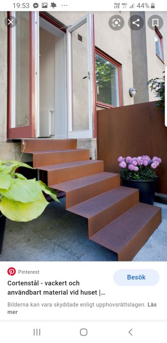 Trappsteg i cortenstål utanför hus med vit dörr och fönster, omgiven av gröna växter och rosa blommor.