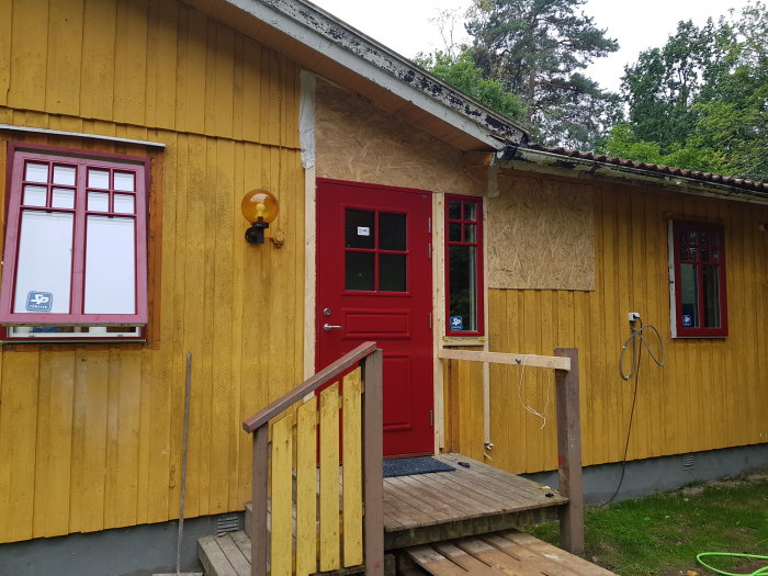 Rött hus med ny ytterdörr och en tilltäppt dörröppning med OSB-skiva på gaveln.