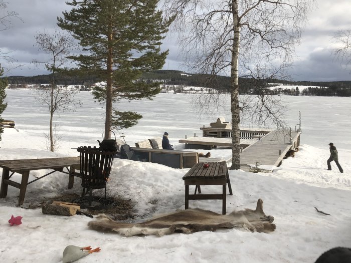 Vinterträdgård med uteplats, eldkorg, brygga och människor vid ett fruset sjölandskap.