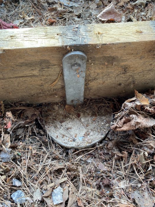 Gammal träbalk stödd på betongplint med fastgjuten plintjärn i ett område med löv och grus.