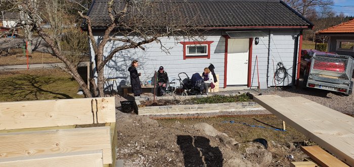 Personer tar en paus från byggprojekt utanför ett hus med byggmaterial i förgrunden.