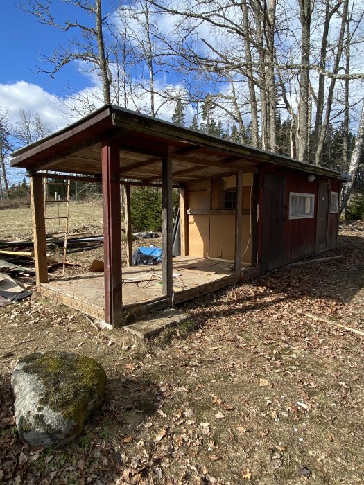 Gammalt förråd halvveis rivet med öppna väggar och synliga stolpar mot en skogsbakgrund under renovering.