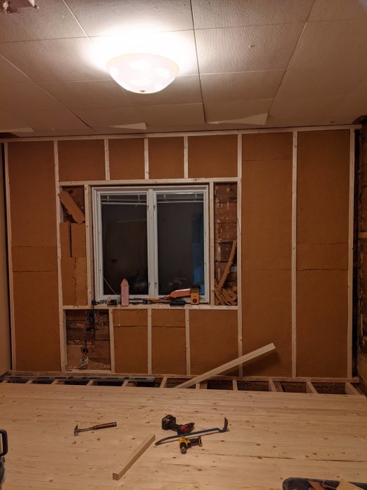 Oisolerade reglar och isolering i en vägg under renovering, med ett fönster, verktyg på golvet och nytt tak.