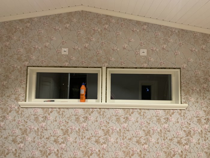 Två nära intilliggande fönster utan foder mellan, på en vägg med blommigt tapetmönster.