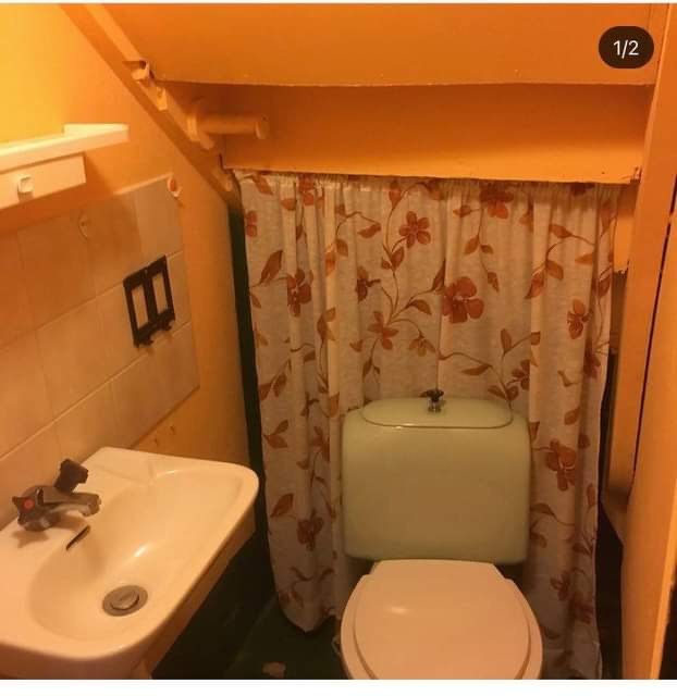 Gammal toalett med orange väggar, tvättställ och toalettstol bakom blommigt draperi.