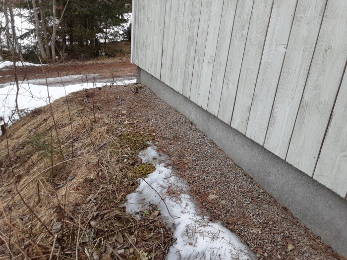 Yttervägg på ett garage med träpanel och betongfundament, intill en snötäckt grusväg och vissnat gräs.