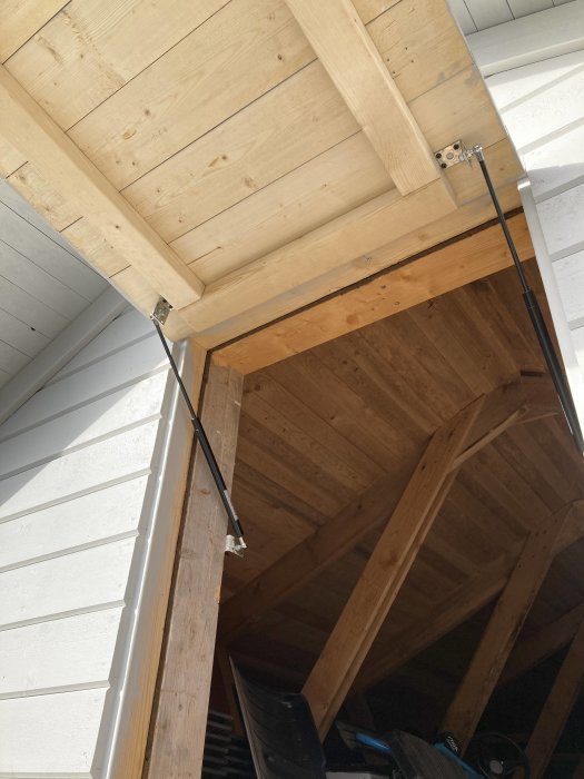 Vindslucka på ett garage med installerade gasfjädrar, öppen och visar träreglar och takkonstruktion.