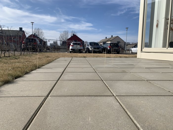 Marksten på uteplats med svagt lutande fall och små ojämnheter, bilparkering och hus i bakgrunden.