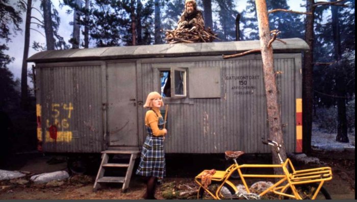 Kvinna står framför en äldre manskapsvagn i skogsmiljö, med en person på taket och en gul cykel framför trappan.