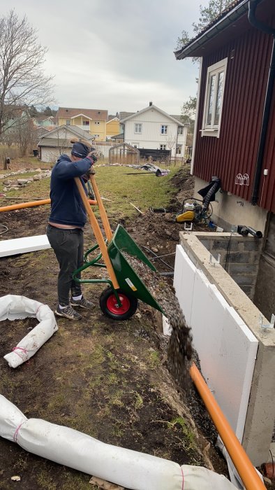 Person som tippa grus från skottkärra vid grund till hus, dräneringsslang och markduk synlig.