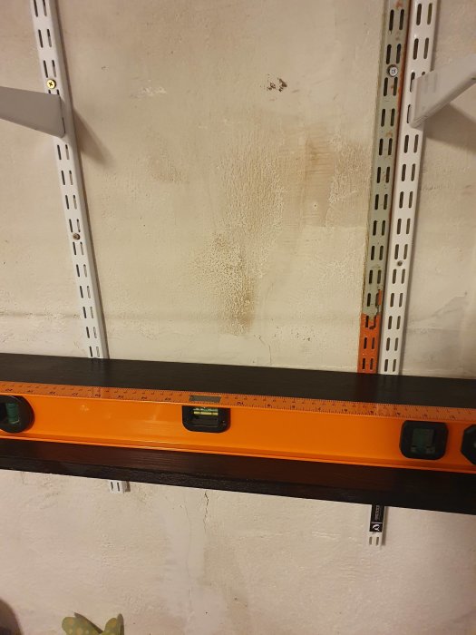 En vattenpass placerat på en hyllplatta i en källare, med synliga väggkonsoler och ojämn bakgrund.