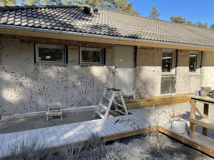 Halvfärdig fasad med borttagen puts och nyligen installerade fönster längs husvägg under renovering.