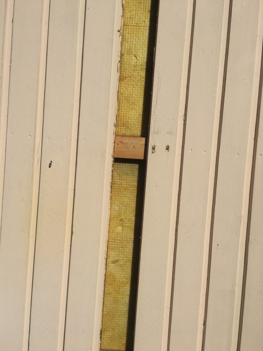 Öppen väggplanka som exponerar gul isolering bakom vitmålade träpaneler på ett hus.