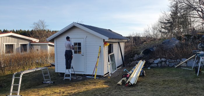 Person står på en stege och arbetar med installation av en ytterdörr på ett litet vitt hus, omgivet av byggmaterial.