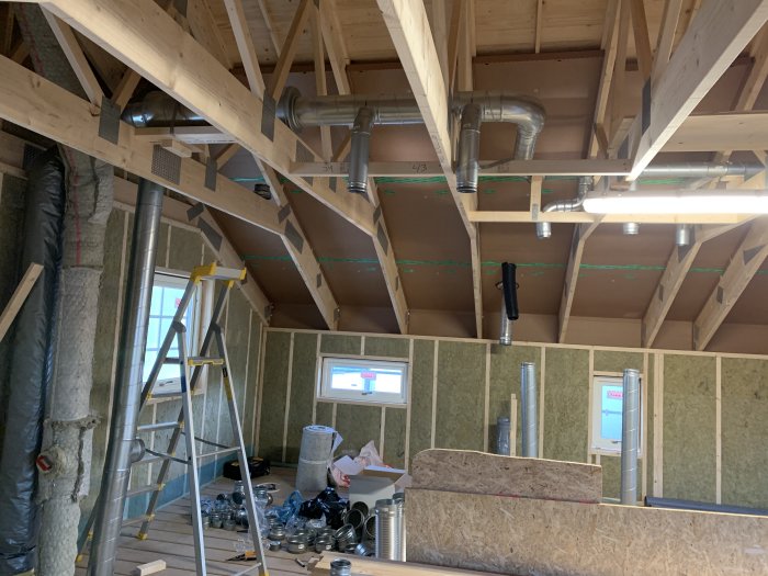 Inredning av hus under konstruktion med ventilationssystem och isolering, stegar och byggmaterial synliga.