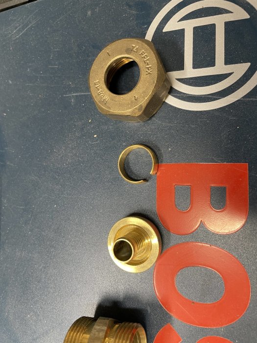 Olika storlekar av mässingmuttrar och packningar på en mörk yta med en röd symbol och delvis synlig text.