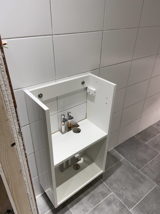 Vit kommod under montering med röranslutningar mot kaklad vägg och grått klinkergolv.