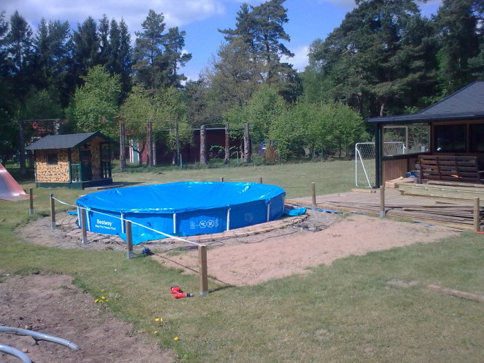 Uppställt blått ovanjordspool med träplankor för ett nedmonterat bjälklag och slang i trädgården.