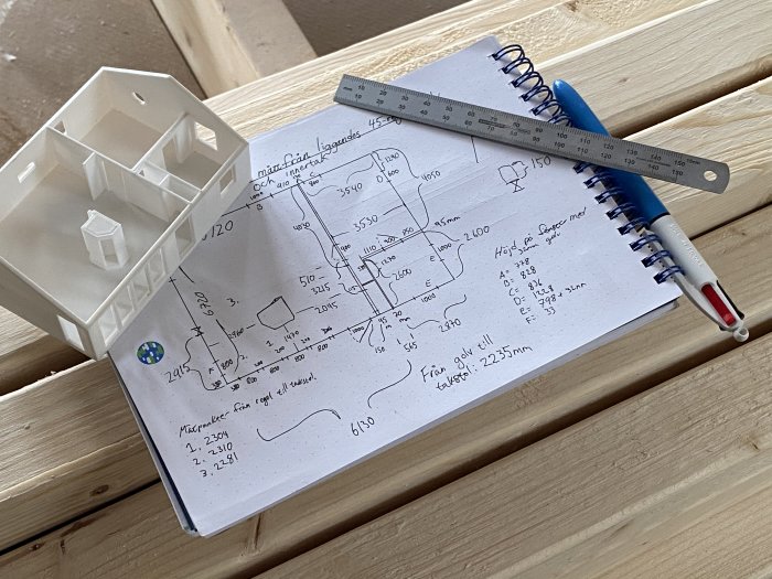 Skrivbok med byggteckningar och mått, linjal och vit 3D-modell av hus på träunderlag.