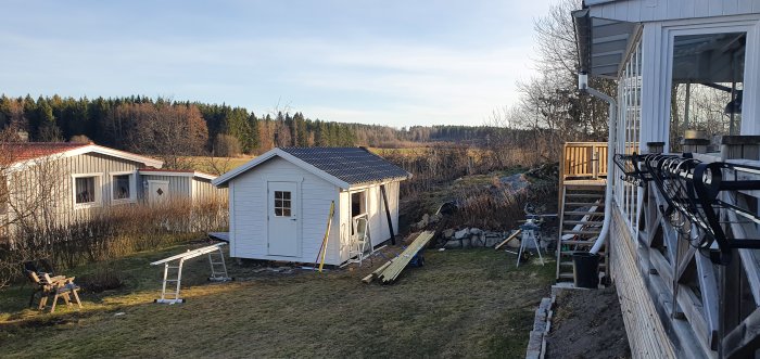 Nyinstallerad ytterdörr på vitmålat uthus med nytt tak och panel, byggarbetsmaterial synligt, i lantlig miljö.