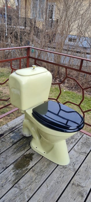 Retro porslinstoalett i ljusgul med svart toalettsits utomhus på en träbrygga.