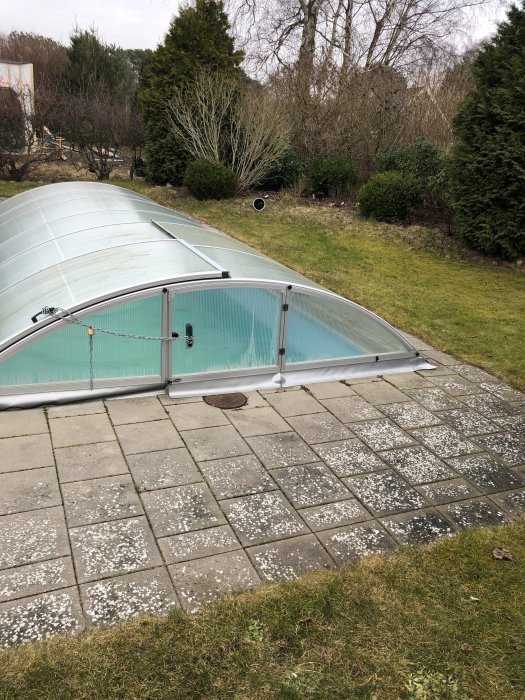 En stenterrass med mönstrade plattor runt en skyddat poolområde, gräs och växtlighet i bakgrunden.