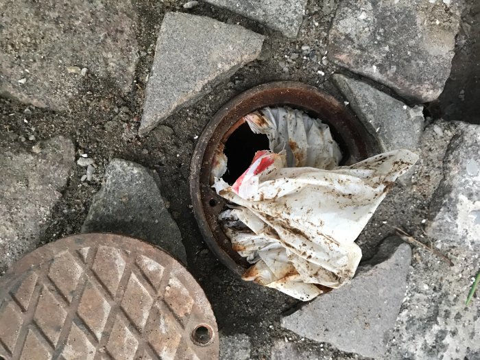 Öppet lock på dränering med keramik-kruka, stenar och trasig plast synlig.