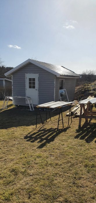 Ett litet gråmålat trädgårdsskjul med öppen dörr, stegar och träplankor för målarprojekt i soligt väder.