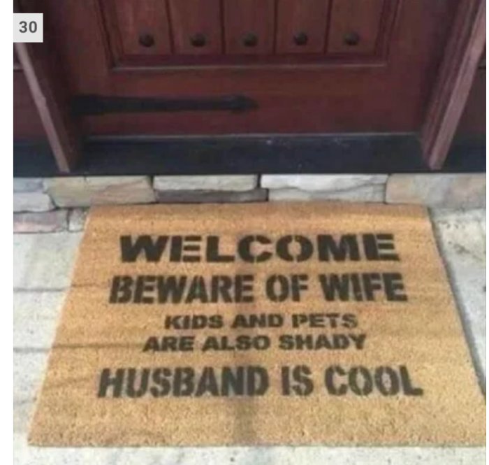 Dörrmatta med text "WELCOME BEWARE OF WIFE KIDS AND PETS ARE ALSO SHADY HUSBAND IS COOL" framför en dörr.