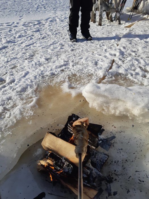 En person som grillar korv över en öppen eld i snölandskap, tydliga vinterspår runtomkring.