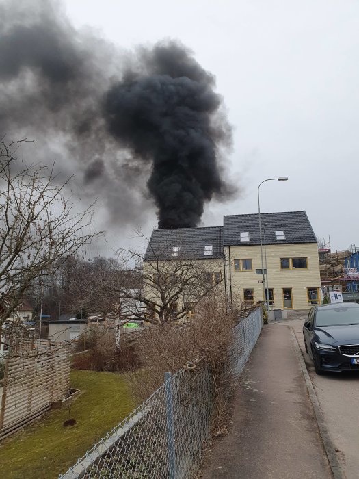 Tät svart rök stiger upp från en byggnad under konstruktion som drabbats av en brand, sedd från en gata i bostadsområde.