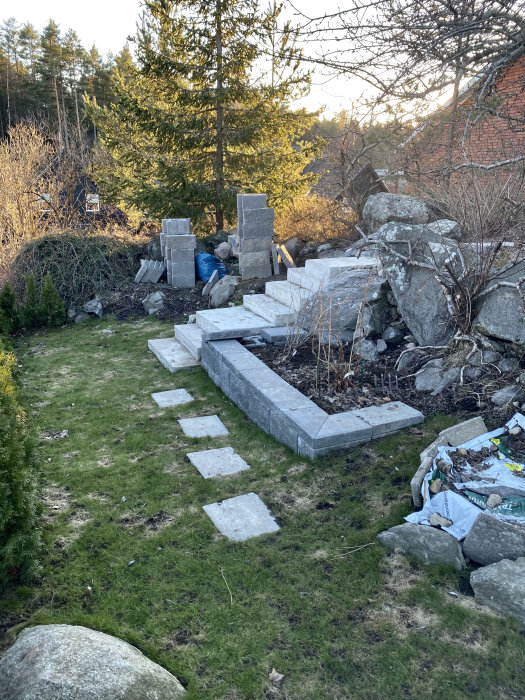 Gräsmatta med kala fläckar och gula partier, stenplattor och oavslutade stenarbeten i en trädgård.