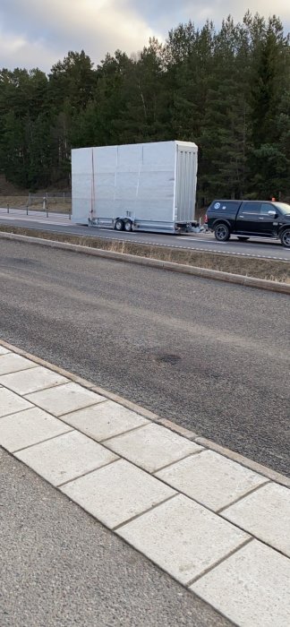 Svart bil som bogserar en stor vit last på en släpvagn på E18 Norrtäljevägen.