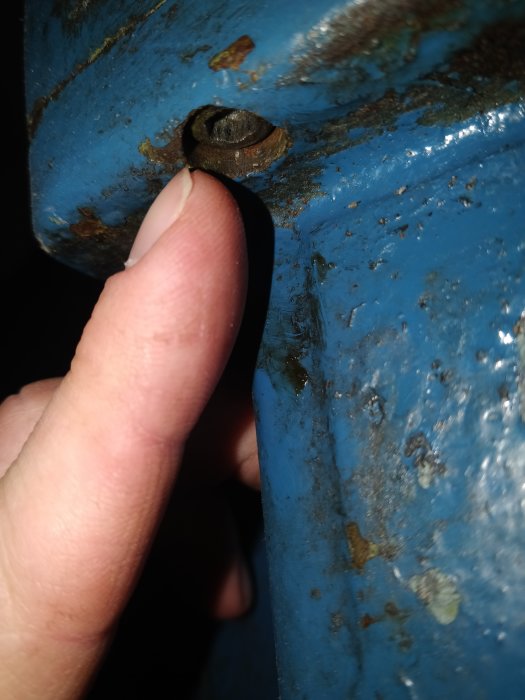 Närbild på en skruv i en blå yta med finger som pekar, smuts och skrapmärken synliga.