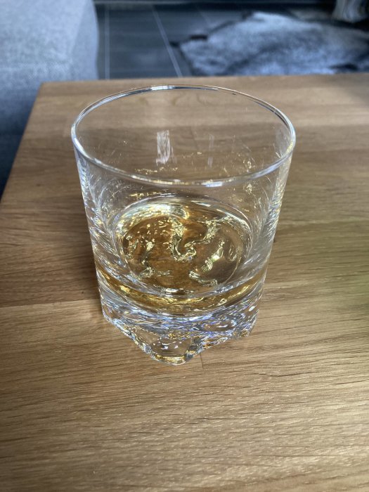 Ett glas med whisky på ett träbord mot suddig bakgrund med grå soffa och ullfilt.