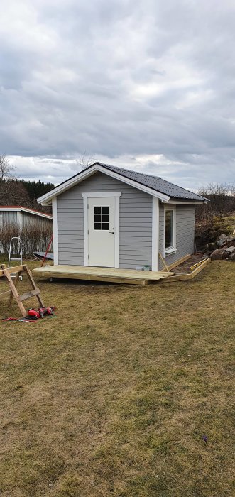 Ett nybyggt grått trädgårdshus med påbörjad altan och löst liggande trallbrädor framför.