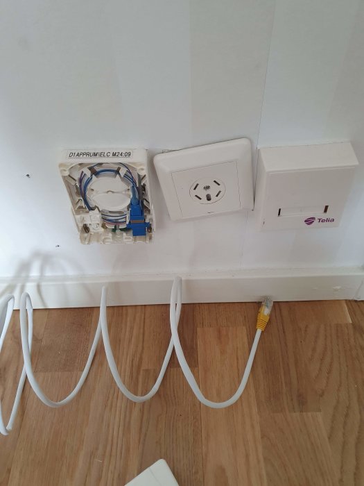 Väggmonterad fiberbox märkt Telia och ofullständig datauttag vid fotlisten, kablar på golvet.
