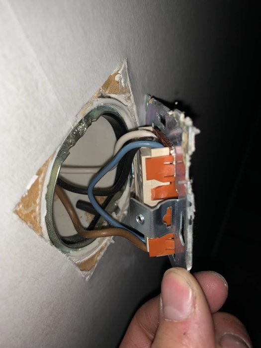 En öppen väggströmbrytare med synliga kopparledningar i svart, vit, brun och blå färg samt orange kopplingsklämmor.