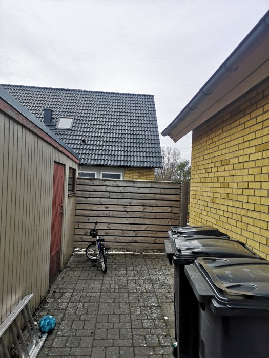 Trång bakgård av ett kedjehus med soptunnor och en cykel, svårtillgängligt för byggarbete.