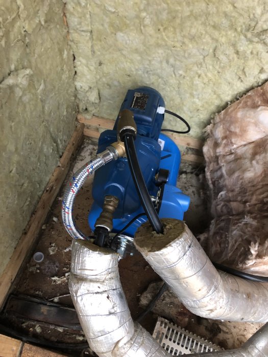 Blå vattenpump installerad i hörnet av ett rum med isolering på väggarna och anslutna sladdar och rör.