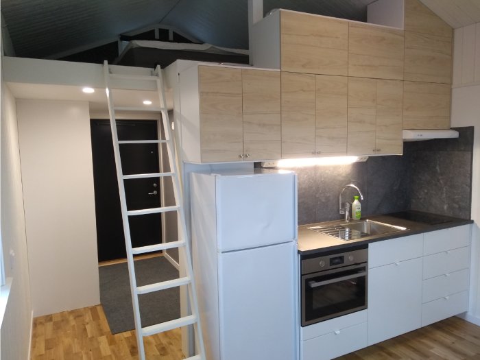 Kök i nybyggt Attefallshus med vita skåp, träbänk, kylskåp, spis och stegar till loftet.