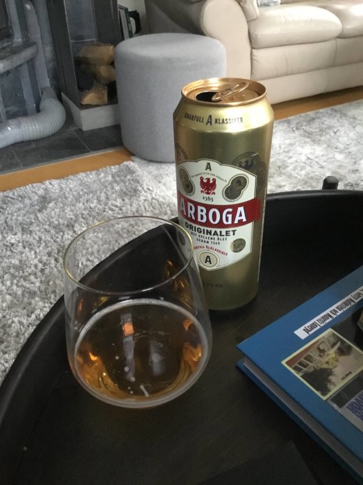 Ölburk av märket Arboga på bord bredvid glas med öl och en soffa i bakgrunden.