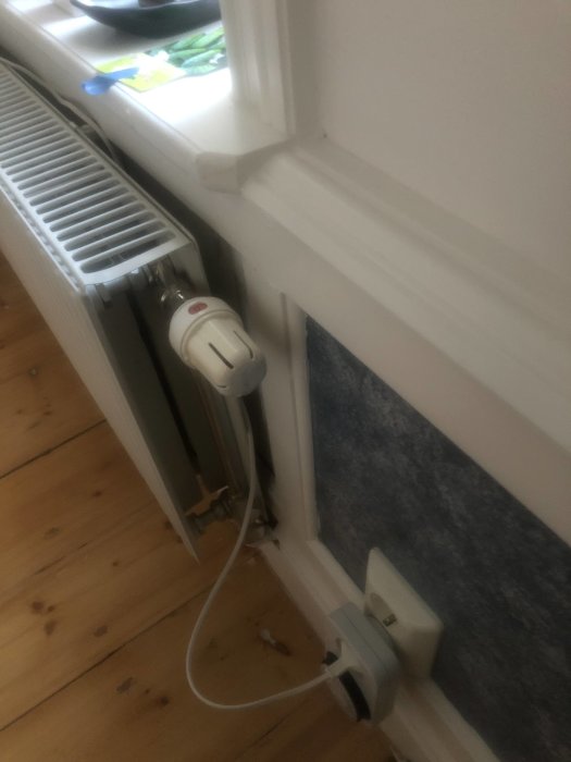 Radiator med termostat nära ett fönster och eluttag i en inomhusmiljö.