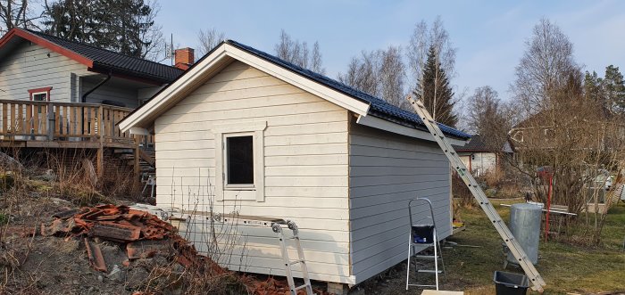 Nybyggt litet hus halvmålat i beige och grått med stegar för målningsarbete.