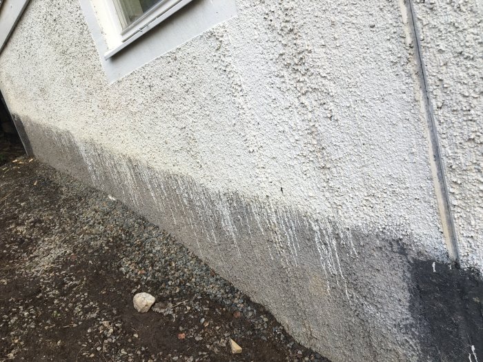 Kortsida av ett hus med ojämn färg och synliga avrinningslinjer på putsad fasad, grus och jord vid grunden.