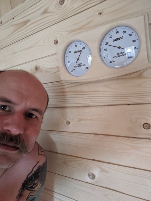 Man halvnaken i bastu med termometer och hygrometer på trävägg.