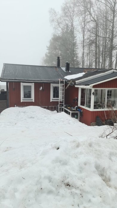 Röd stuga med snedtak och utstickande del under renovering i vinterlandskap, stege lutad mot fasaden.
