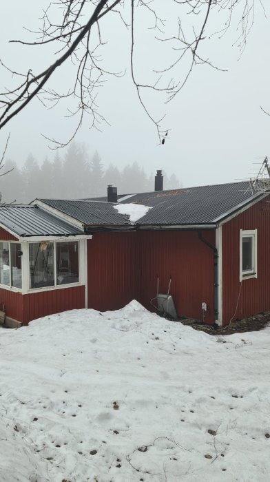 Röd stuga med brutet tak och inglasad farstukvist, omgiven av snötäckt mark och dimma.