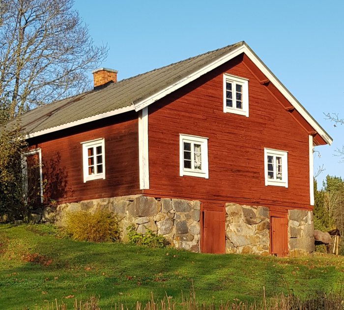 Rödmålad gammal drängstuga från 1850 med vitkantade fönster och en stenfot på en solig dag.