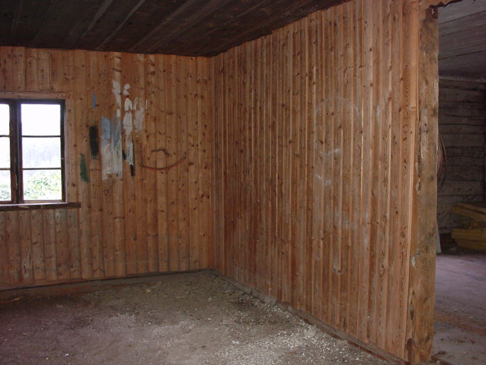 Ett avskalat rum med träpanel på väggarna och synligt timmer, jordgolv, utan inredning och spår av rivningsarbete.